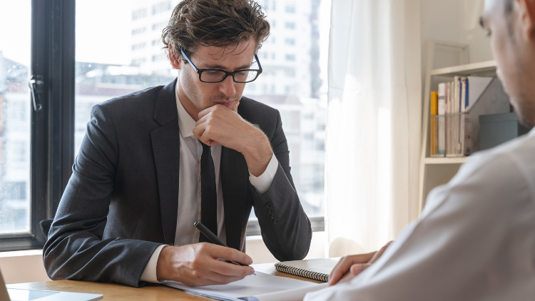 A man wearing a business suit and a pair of glasses looks and reads a business document showed by another man.