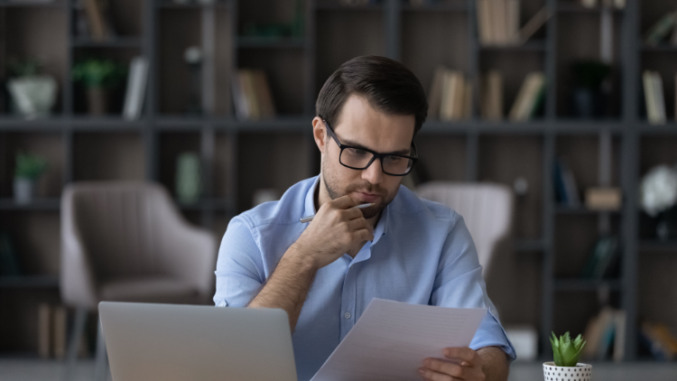 In a home office, a man wearing a blue shirt and glasses reads through a document and contemplates the best next step to grow his business.