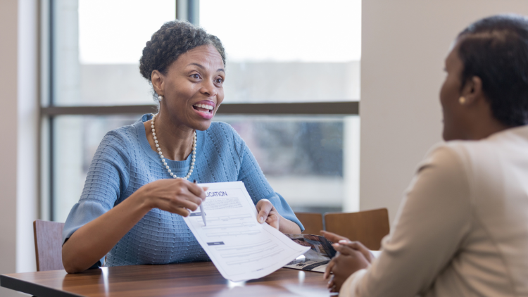 A professional woman in a blue dress smiles while holding and explaining a business account application form to another woman across the table in a bright office setting.