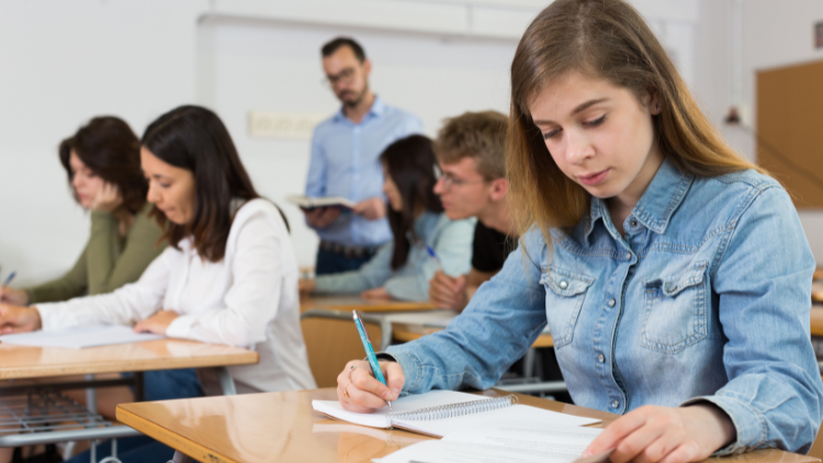 In a classroom, a young girl wearing a denim shirt is looking at a worksheet and writes answers on her notebook.