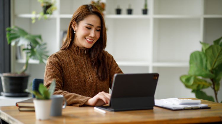 A young lady smiles while working on her tablet set on a home office table.