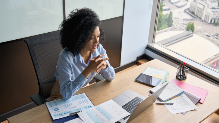 A young professional woman sits at her office desk by a large window, reviewing financial documents and charts spread out beside her laptop while focusing intently on the screen.