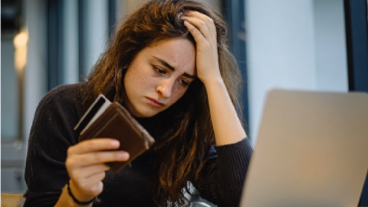 A young woman holding her wallet and credit card while sitting in front of her laptop looking dejected.