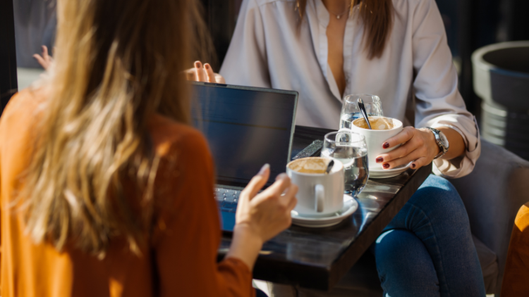Two women sit at a small café table with laptops and coffee cups, discussing work while enjoying a casual business meeting in a relaxed setting.