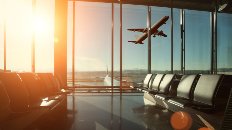 An airplane takes off at sunset, viewed through large glass windows from empty airport terminal seating.