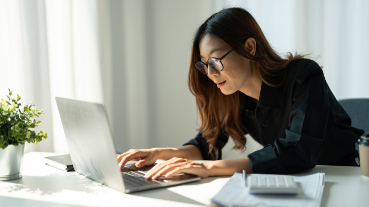 An Asian small business owner wearing glasses works intently on a laptop at a bright home office desk, with documents, a calculator, and a coffee cup nearby.