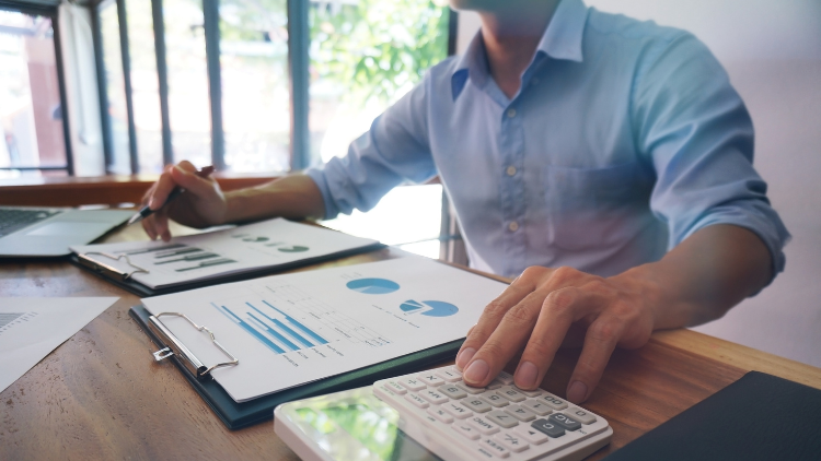 A man in a blue shirt reviews financial charts and uses a calculator while working at a desk with sunlight coming through the window.
