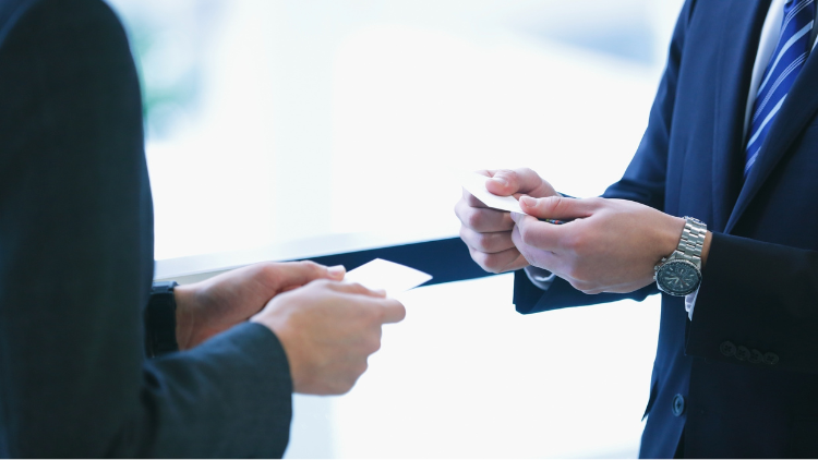Two businessmen in suits exchange business cards in a professional setting, highlighting networking and business relationships.