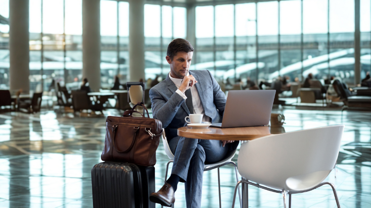 A businessman in a suit works on his laptop at an airport café table with luggage beside him.