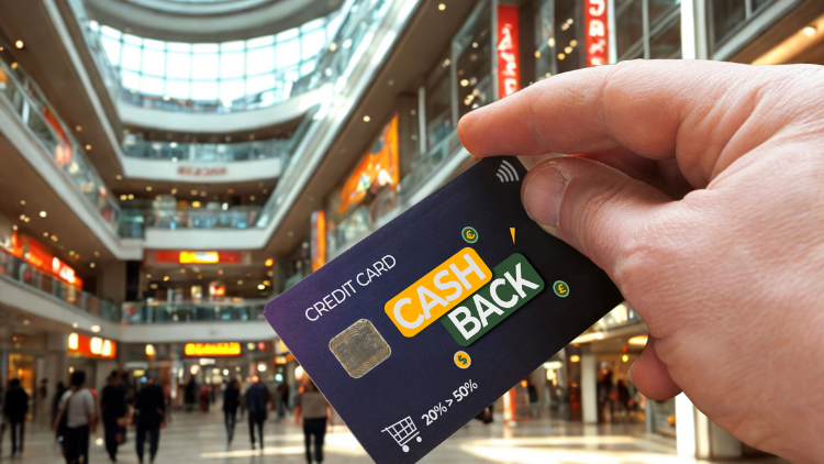 A hand holds a cashback credit card inside a busy shopping mall with stores and people in the background.