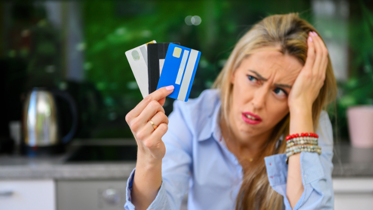 A visibly frustrated woman with her hand on her head holds up three different credit cards, looking confused.