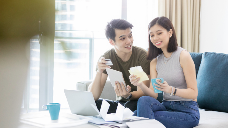 A smiling couple sits on a sofa using a laptop and paperwork to plan household finances.