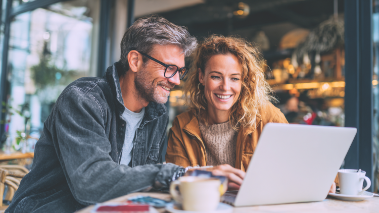 A smiling couple sits together at a café table, using a laptop to plan their finances and rewards strategy.