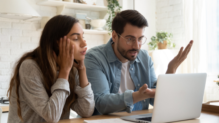 A young couple sits at a table looking frustrated while reviewing financial documents on a laptop.