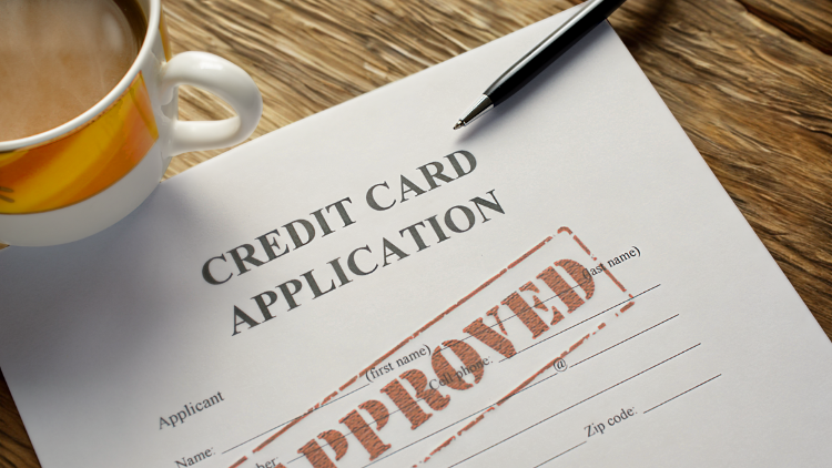 A close-up of a credit card application form on a wooden table with a coffee cup and pen, stamped “approved.”