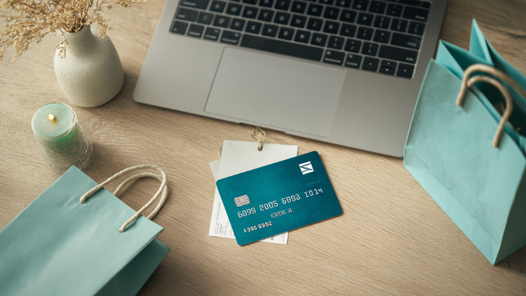A credit card is placed on a desk with shopping bags beside a laptop.