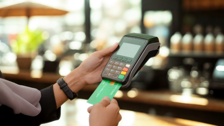 A person swipes a green credit card through a payment terminal at a store counter.