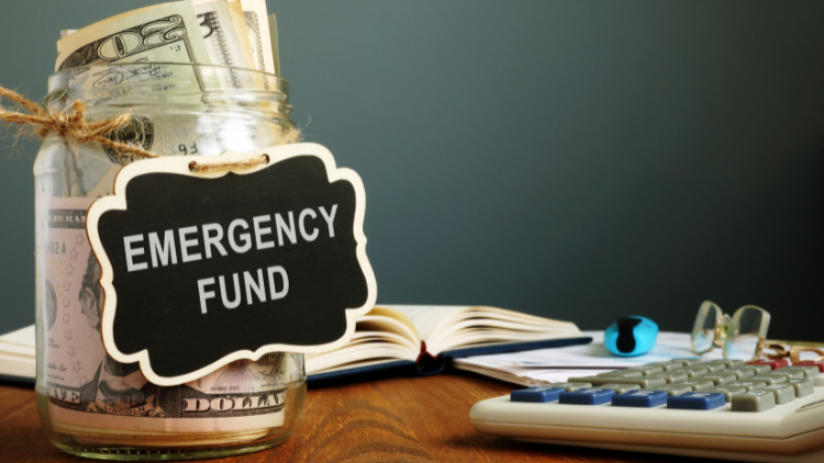 A glass jar labeled emergency fund filled with dollar bills sits on a desk beside a calculator and notebook.