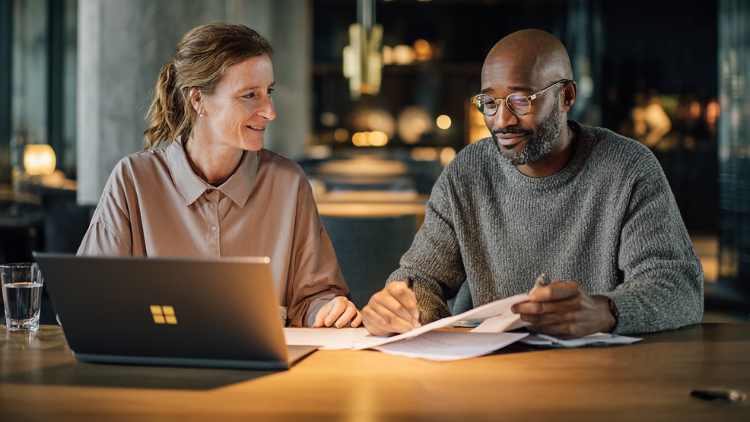A man and woman sit at a table with a laptop and paperwork while discussing finances together.