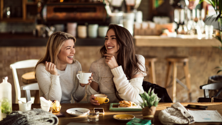 Two women laughing together over coffee and pastries in a café, showing how casual meals can still earn dining rewards points.