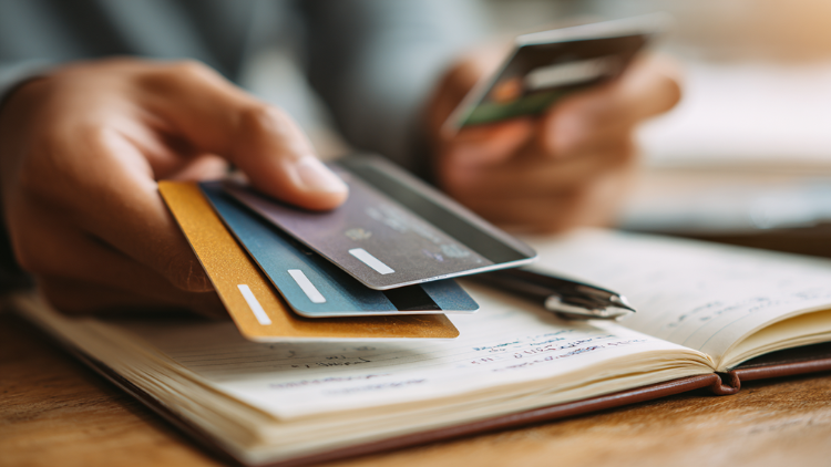 A person’s hand holds three credit cards over a notebook and a pen, appearing to decide which one to use.