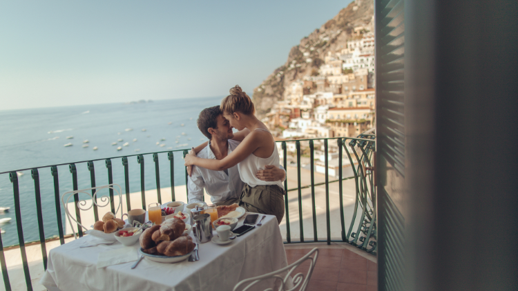 A couple shares a kiss during breakfast on a balcony overlooking the ocean and coastal town.