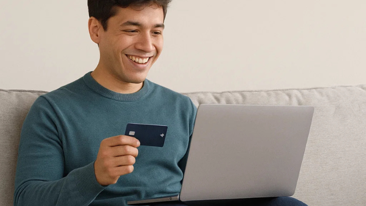 Smiling man sitting on a couch holding a credit card while shopping online with his laptop.