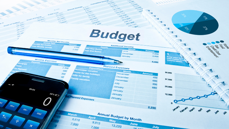 Financial budget documents with charts, graphs, a calculator, and a pen are spread across a desk for monthly expense planning.