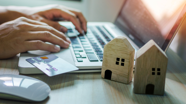 Hands typing on a laptop with a credit card and two small wooden house models on the desk.