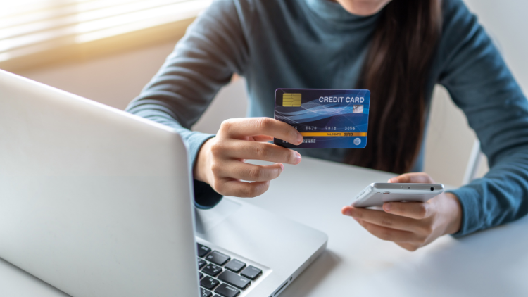 A person holds a credit card in one hand and a smartphone in the other while shopping online at a laptop.