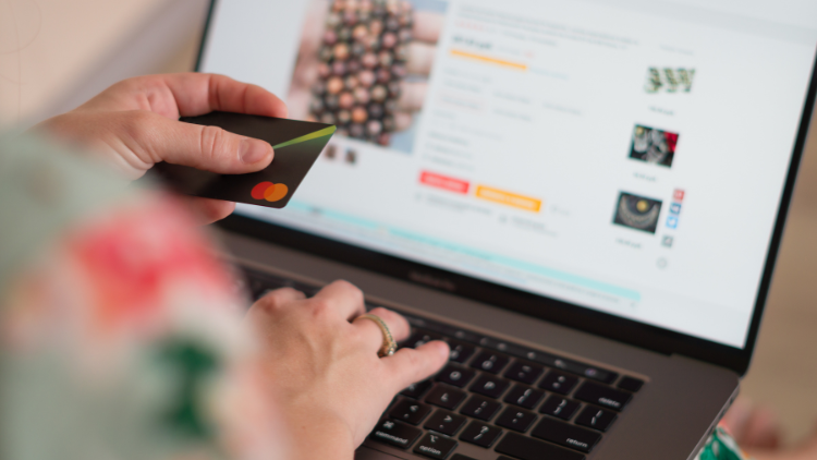 A person holds a credit card while shopping for jewelry online using a laptop.