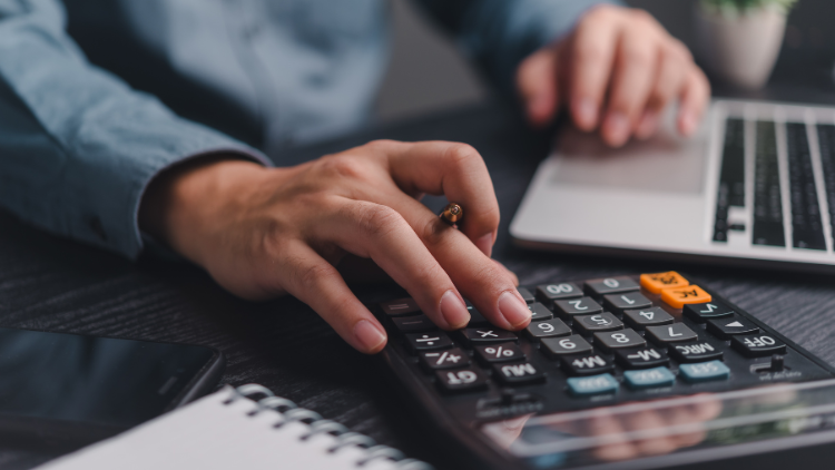 A person presses buttons on a calculator while working on a laptop at a desk with a notepad and smartphone.