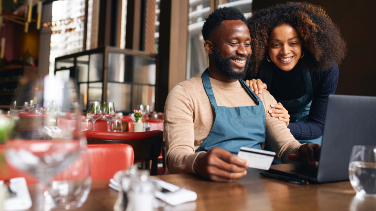 A smiling couple wearing aprons sit in a restaurant using a credit card and laptop together for business expenses.