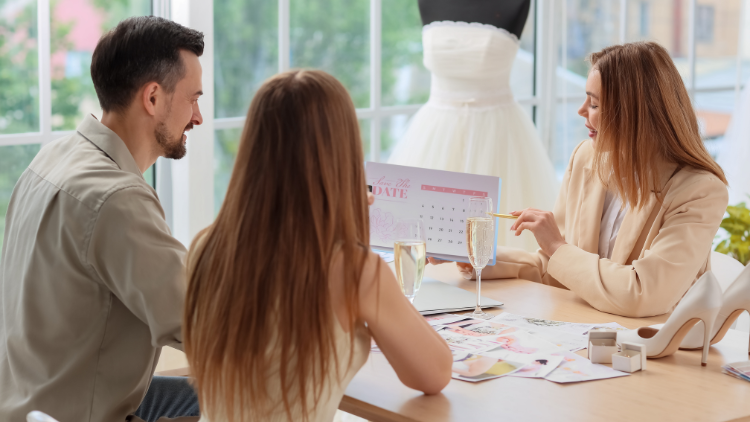 A bride and groom consult with a wedding planner reviewing a calendar and bridal details at a table.