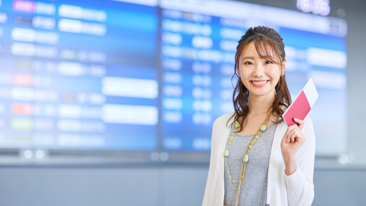 A smiling traveler holds her boarding pass at the airport in front of a flight information display board.