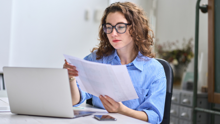 Woman in glasses reviewing bills while working on laptop.