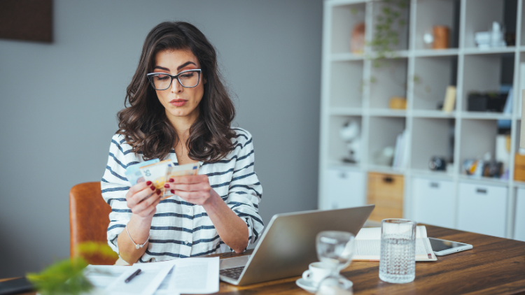 A woman in glasses sits at a desk with a laptop and papers while carefully counting cash.