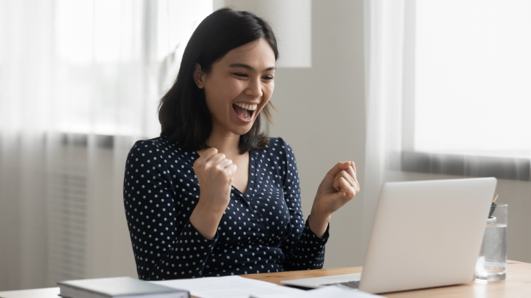 A smiling woman raises her fists in excitement while looking at her laptop screen at home.
