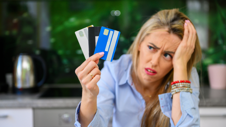 A frustrated woman holds multiple credit cards in her hand while sitting in a kitchen, showing concern about financial choices.