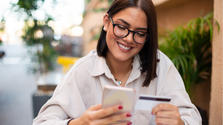 A smiling woman with glasses uses her smartphone while holding a credit card at an outdoor café.