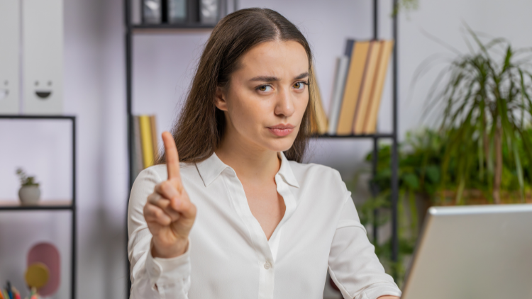 A serious woman in a white blouse raises her index finger in caution while sitting at a desk.