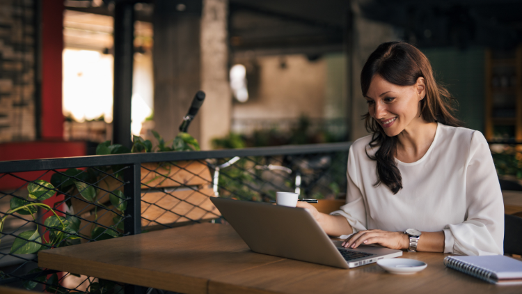 A smiling woman works on her laptop while drinking coffee at a café table.