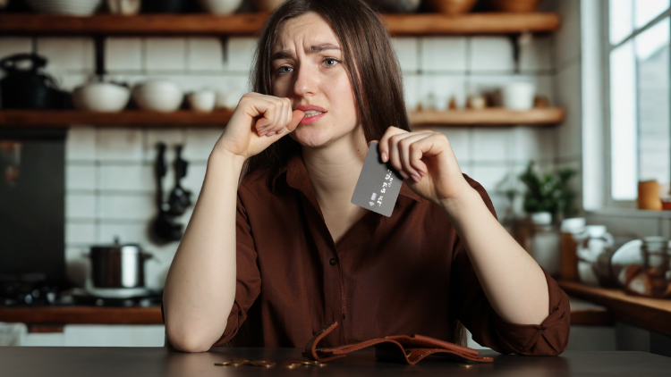 A concerned woman sits at a kitchen table holding a credit card with a nearly empty wallet in front of her, reflecting financial stress.